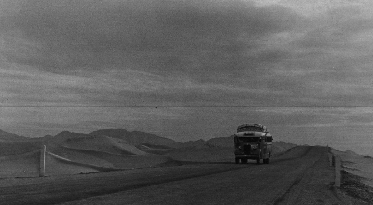 Black-and-white photograph of a passenger bus driving through the desert.