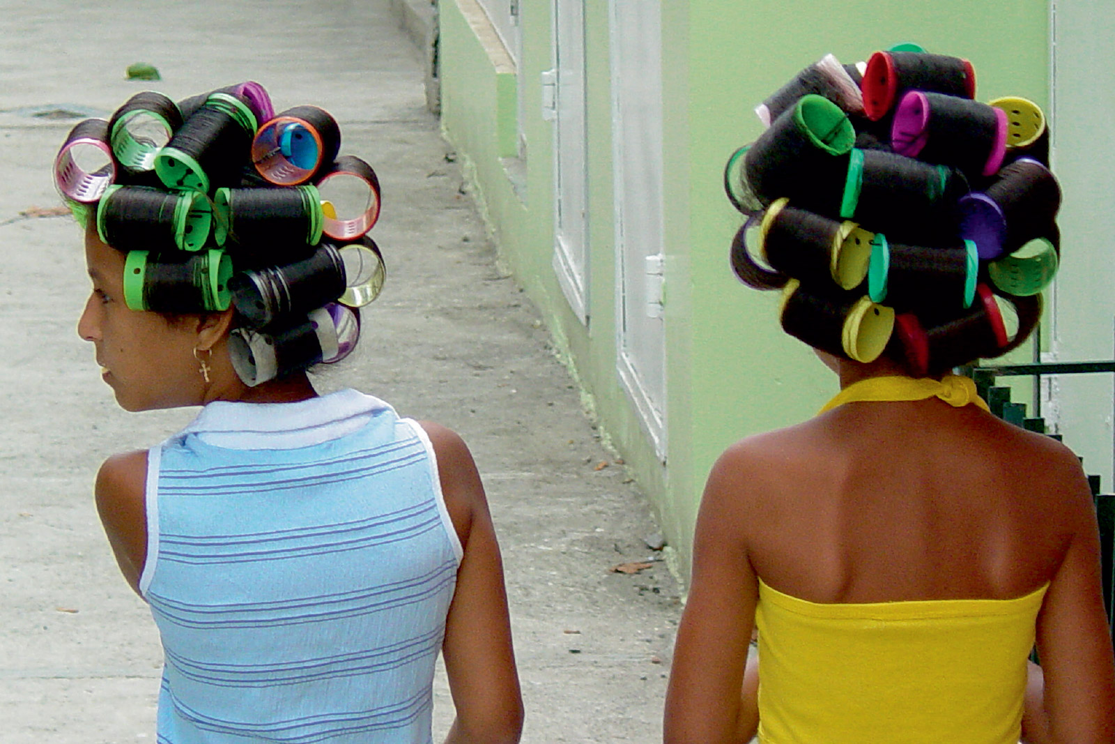 A postcard of a two thousand four photograph by Adam Jones, showing two women from behind wearing colorful hair curlers in San Jose de Ocoa. 