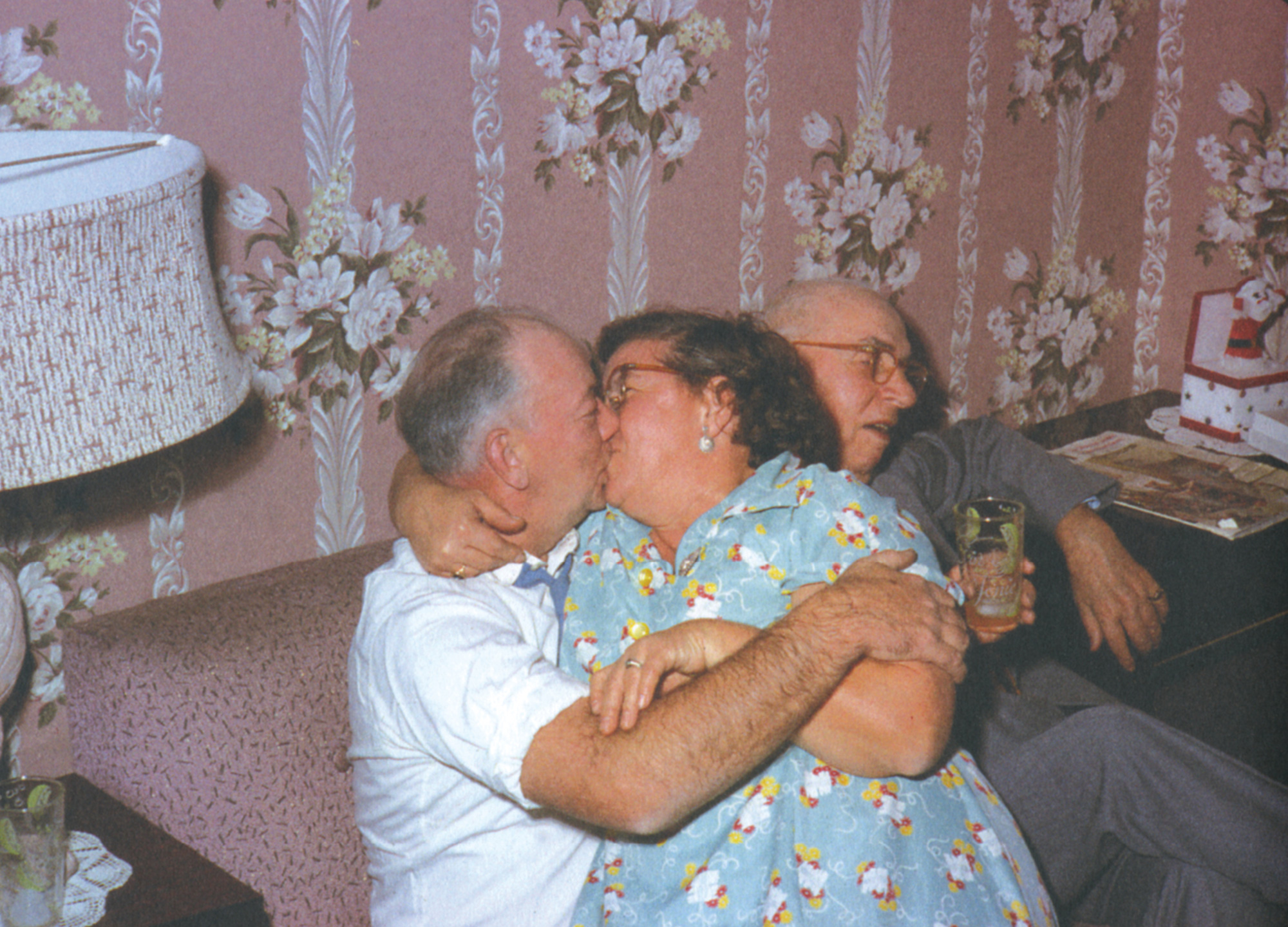 A photograph of an elderly man sitting on a couch, almost crushed by a couple who are passionately kissing directly beside him.
