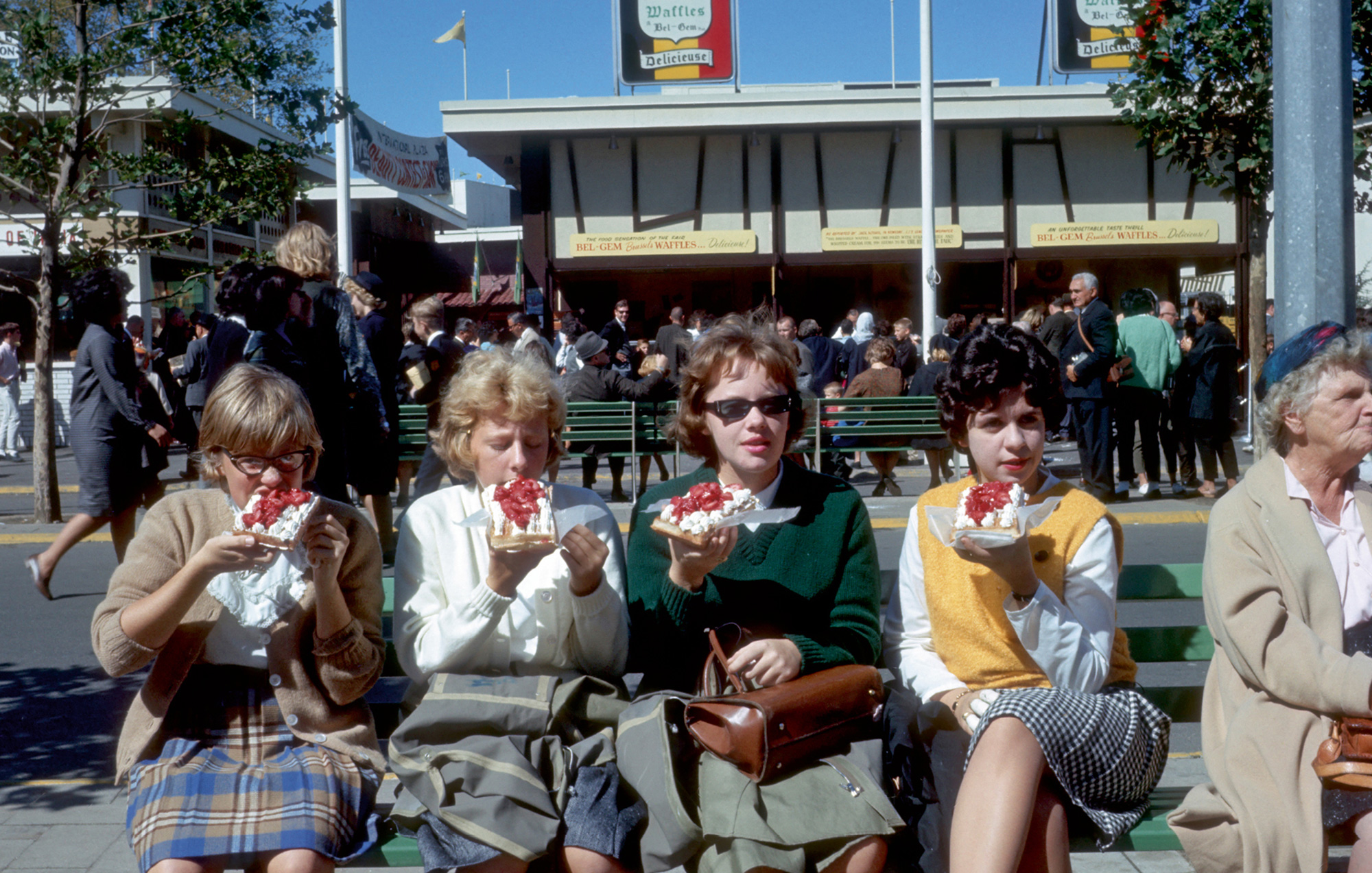 A photograph of four women eating Bel-Gem waffles at the nineteen sixty-four World’s Fair in New York. The snack proved so popular that the Vermersch family opened a number of additional stands during the course of the fair, such as this one in an area known as the International Plaza.