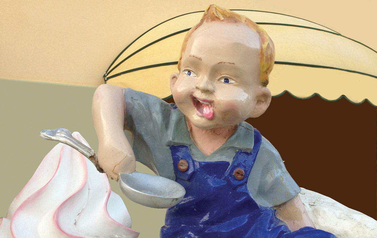 A photograph of Melanie Jackson’s twenty sixteen fiberglass sculpture “The Lick Two,” of a young boy holding a spoon with his tongue out. This mascot from northern France was part of a larger display advertising ice cream.  