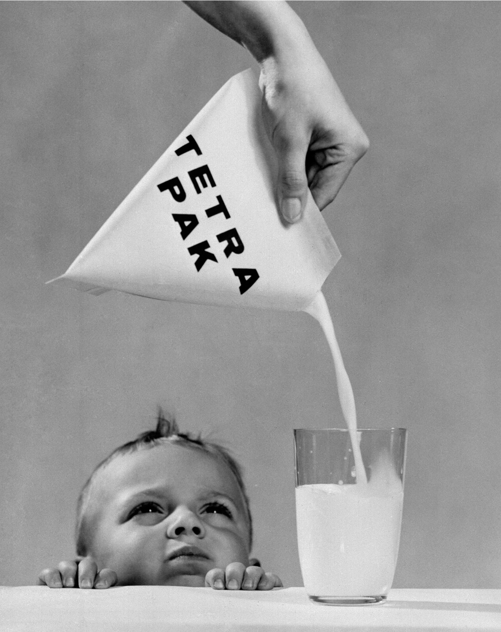 A nineteen fifties promotional image for Tetra Pak, featuring a baby gazing at a glass of milk being poured from a packet.