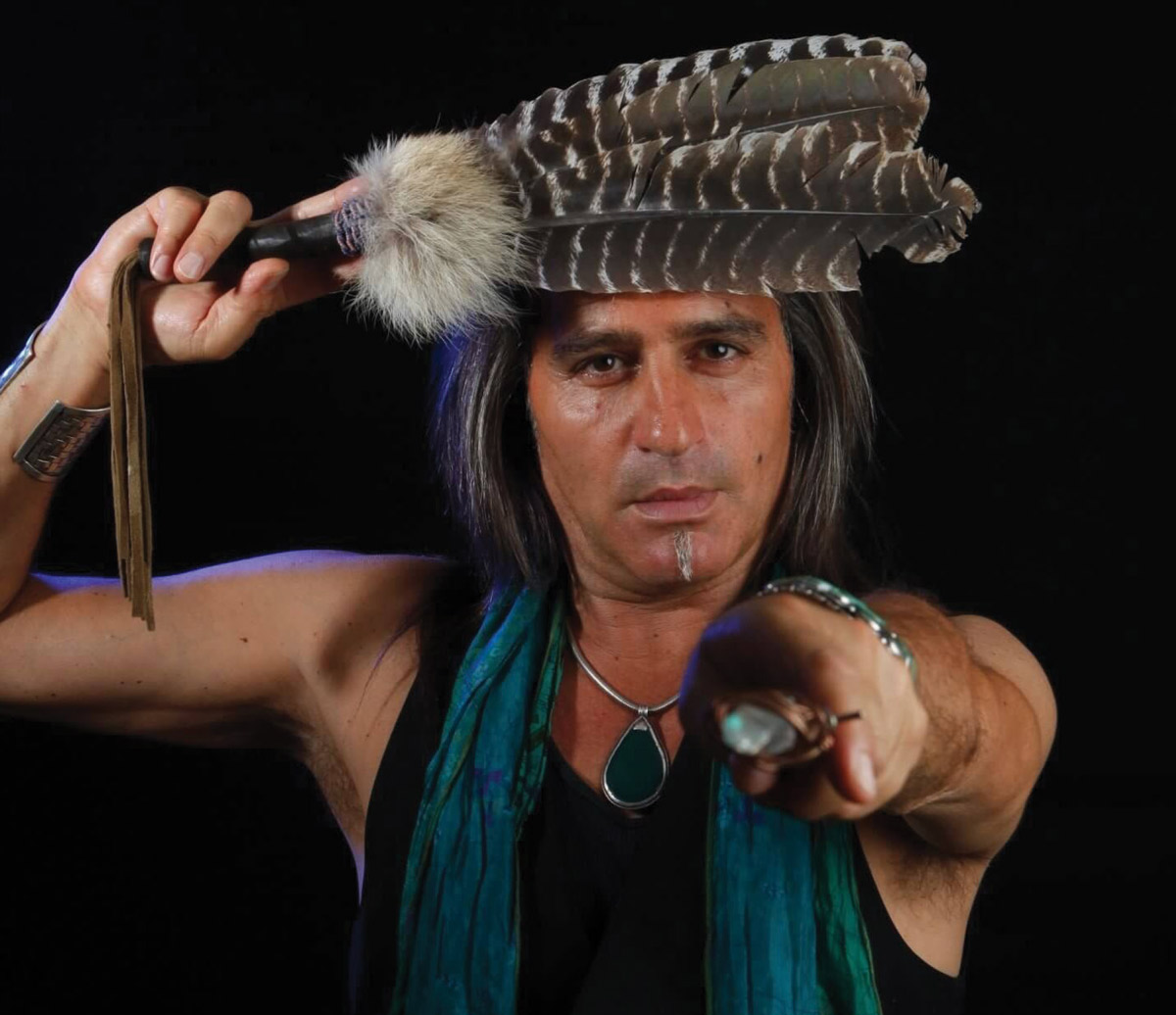 A photograph of a man posing in costume Native American apparrel and accessories. 