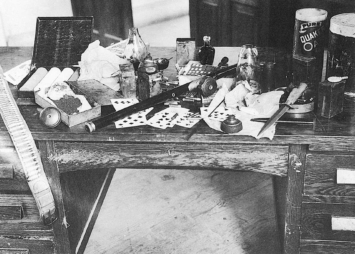 A photograph from the 1920s or 1930s depicting a desk covered with items including playing cards, a knife, and drug paraphernalia. 
