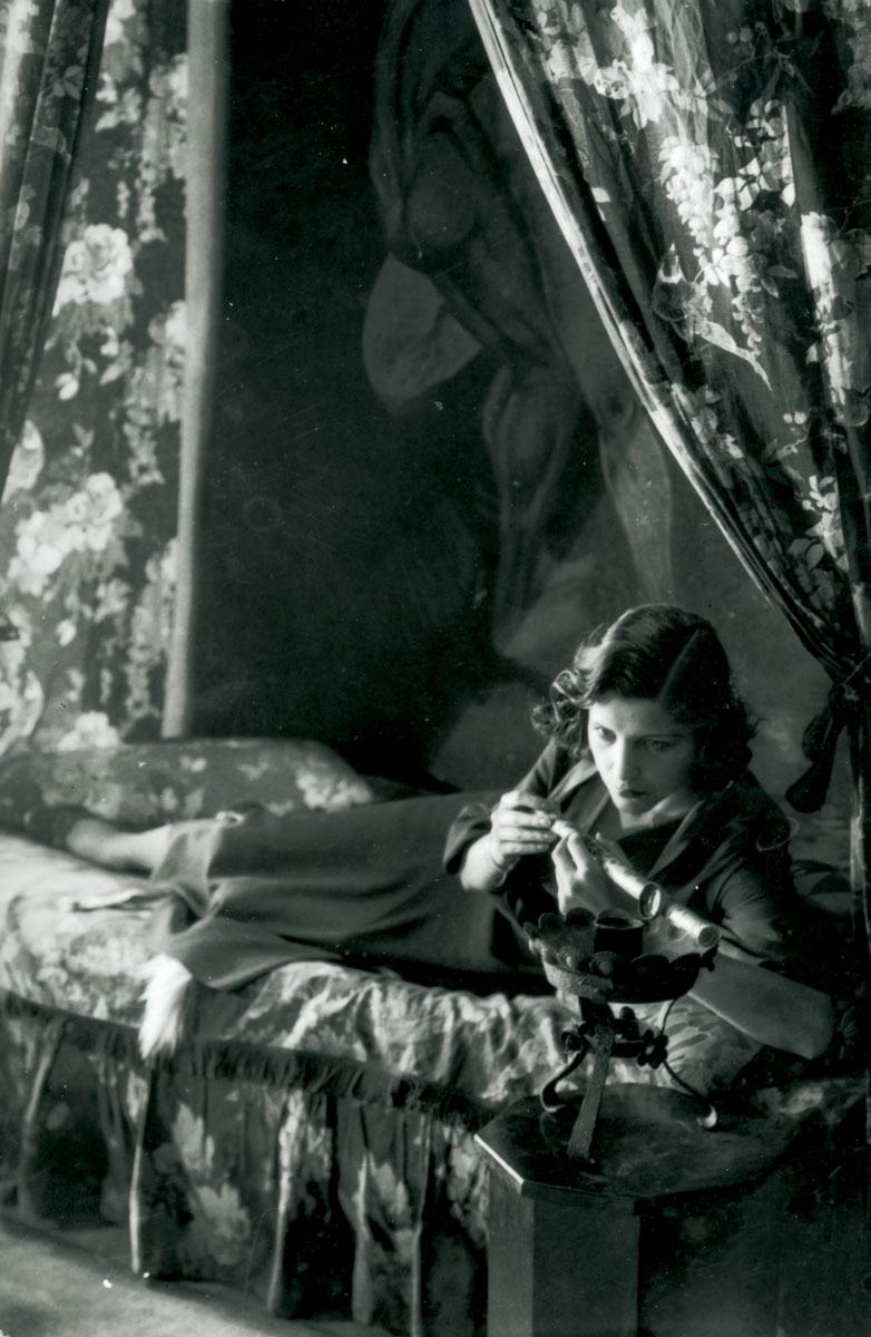 A photograph from the 1930s depicting a woman lying on a bed and holding an opium pipe.