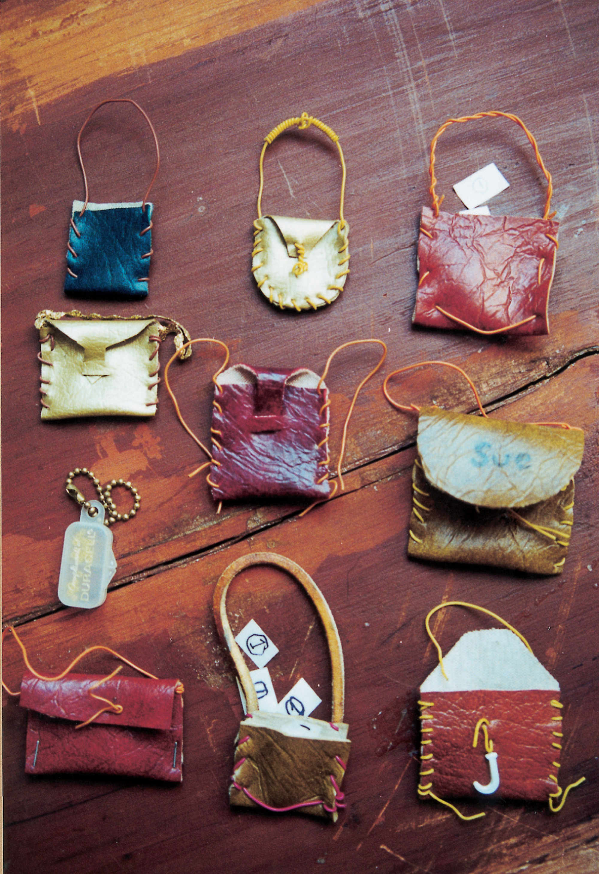 A photograph by of a number of small crafted bags. The caption reads: Eight assorted handbags, one purse and one backpack. The Duracell purse, semi-transparent purse on small brass bead-chain, was made from a container for hearing aid batteries. All others are of faux leather, most stitched with telephone wire. Two contain paper “money,” white rectangles each bearing a circled numeral 1 in pencil/black marker. Paradoxically, although the Doll Games showed a utopian disregard for money and a high scorn for the conventional appurtenances of femininity, purses were manufactured in quantities rivaled only by daggers. Was the vaginal purse—pictured here in an almost military array—waging a war with the phallic dagger over the contested territory of the Doll Games?
