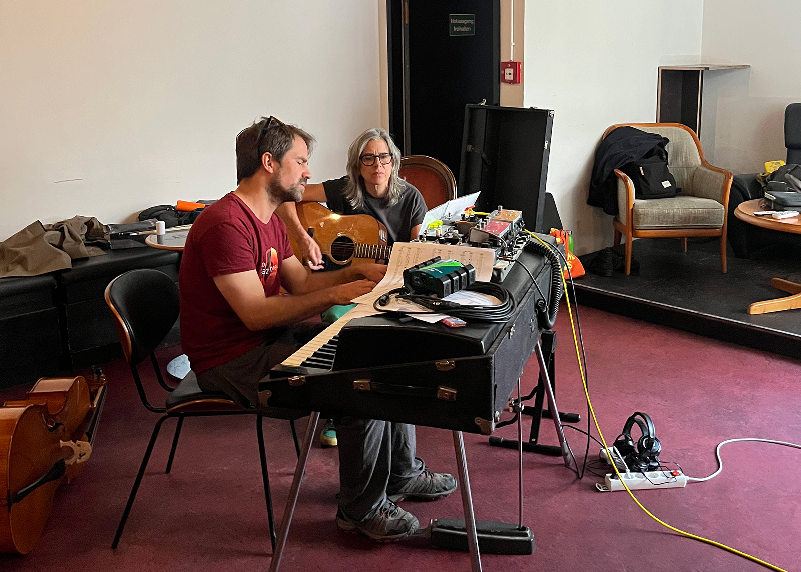 A photograph of a performer sitting before a keyboard and another performer holding a guitar as they rehearse a duet together.