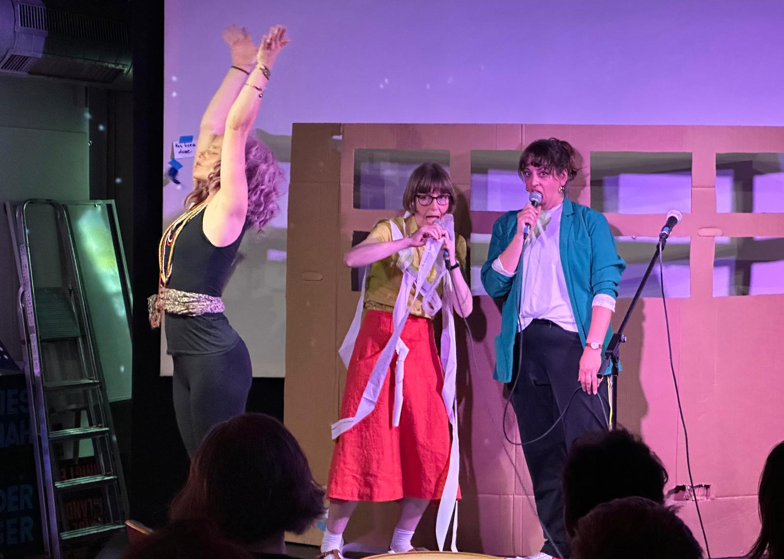 A photograph of one performer raising her arms as she demonstrates a yoga-inspired breathing ritual and two other performers breathing exaggeratedly into their microphones.