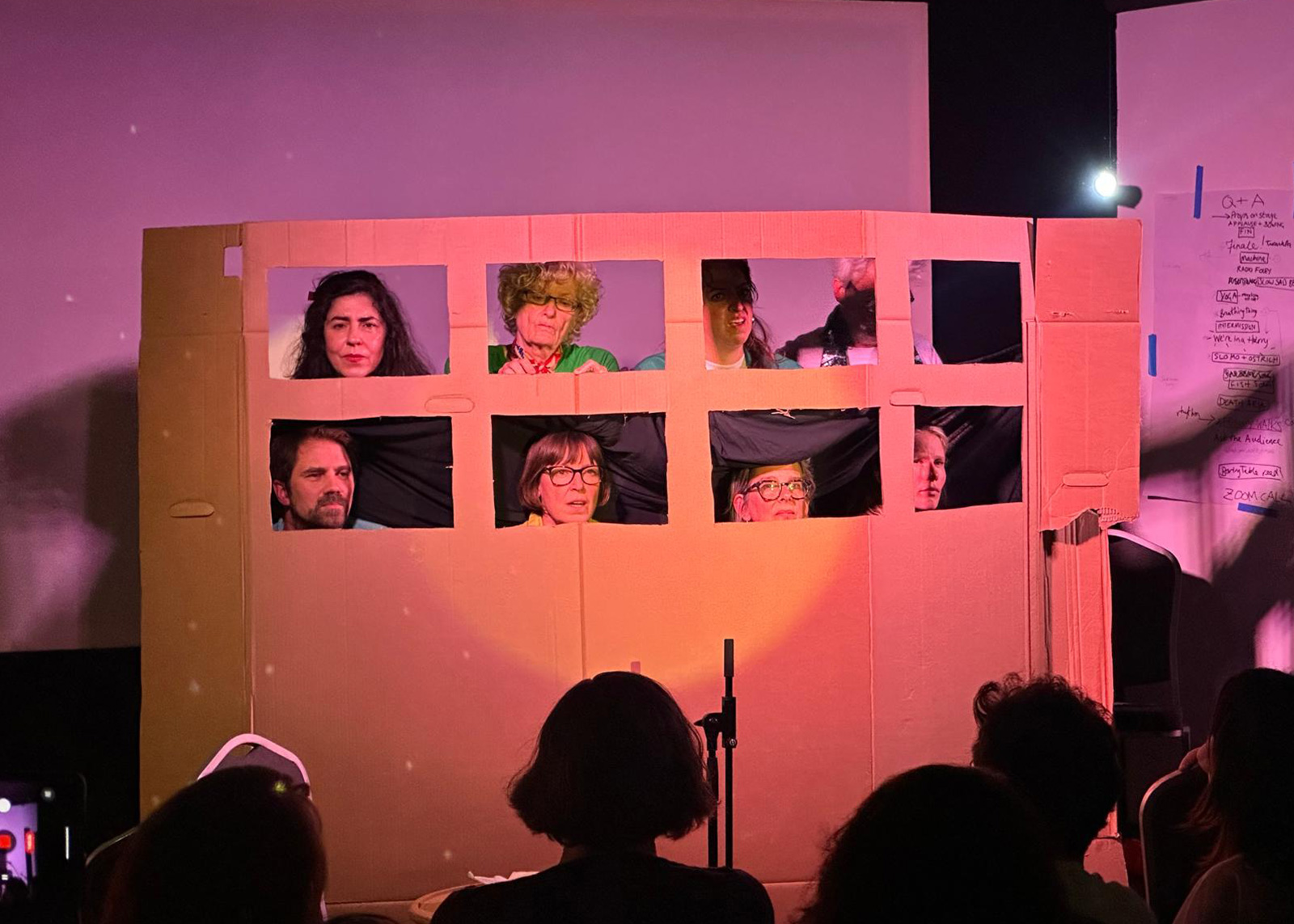 A photograph of the performers enacting a Zoom call by positioning themselves behind a sheet of cardboard that resembles a computer screen, with cut-out squares for their faces.