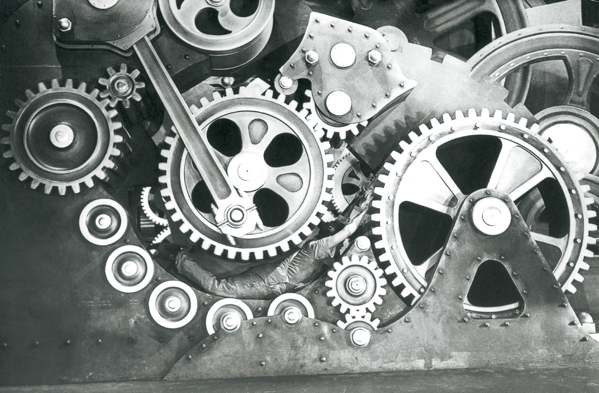 A film still from Modern Times depicting Charlie Chaplin getting stuck in the cogs of a machine.