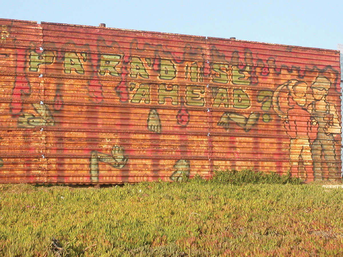 A photograph of a section of border fence painted with the question “paradise ahead?”