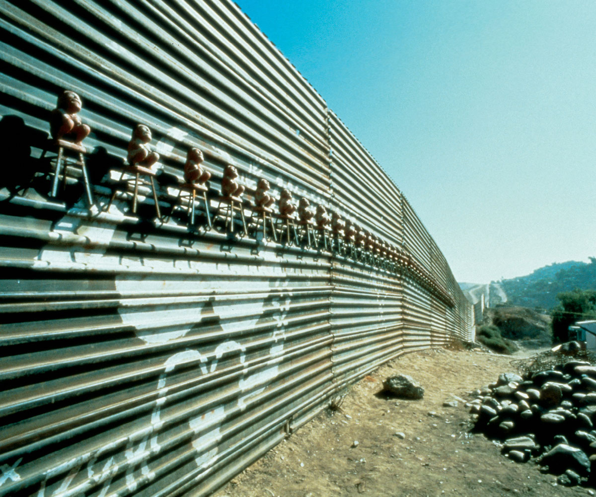 A photograph of a 1994 mixed media installation of small figures sitting on the border fence by Silvia Gruner, entitled “The Middle of the Road / La mitad del camino.”