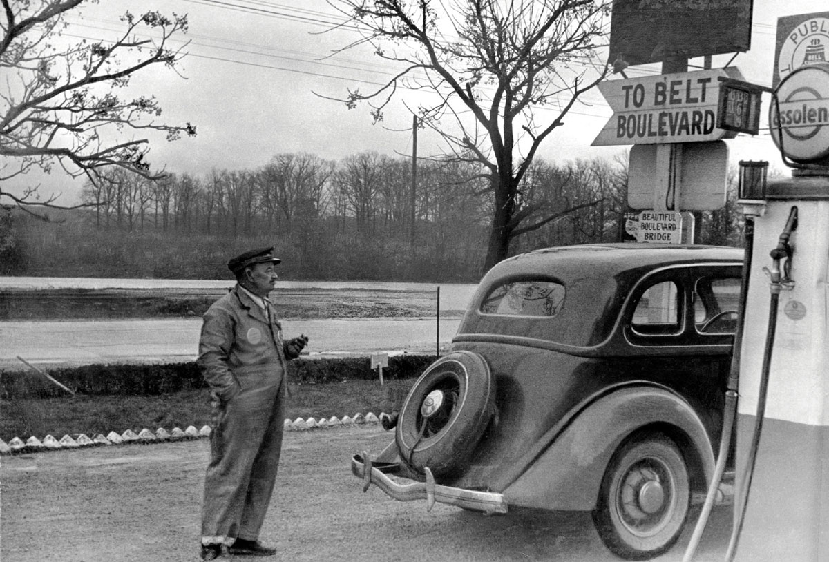 Black-and-white photograph of a gas station attendant standing near a car.