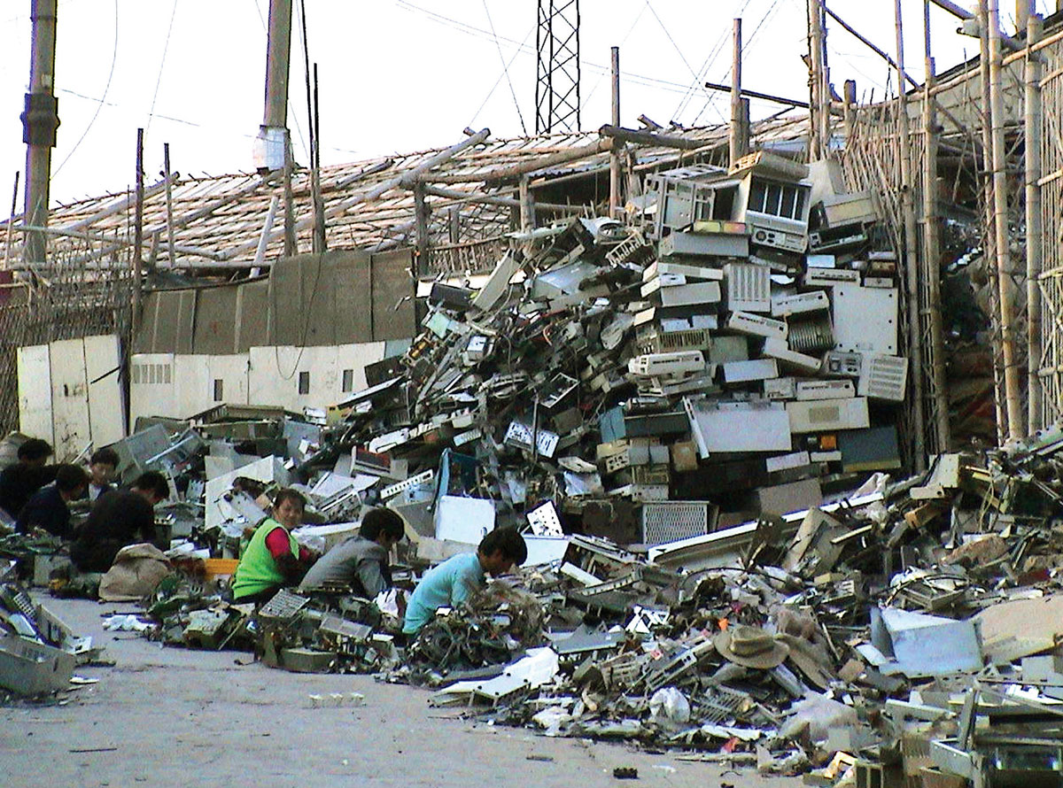 A photograph showing the bleak work conditions of a Chinese scrapyard as workers squat by towering piles of e-waste.