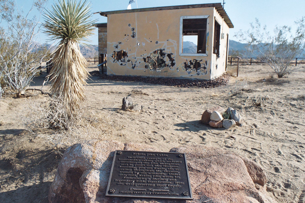 Krblin Jihn Kabin, a Kymaerica historical site in the nation of Notgeon (near Joshua Tree, California). The historical marker in the foreground reads “Well-sited example of the homestedler cabins which secluded members of the Jihn Wranglican sect in post-Civil War Notgeon. Kneeling, you see Kwale, the so-called Wranglican Mt. Hermon, framed by the windows. Under this roof, Krblin Jihn wrote his Kmmentaries n Matthew and legendary Bible translations. Note the well-preserved floor kmpass.” Photo Eames Demetrios.
