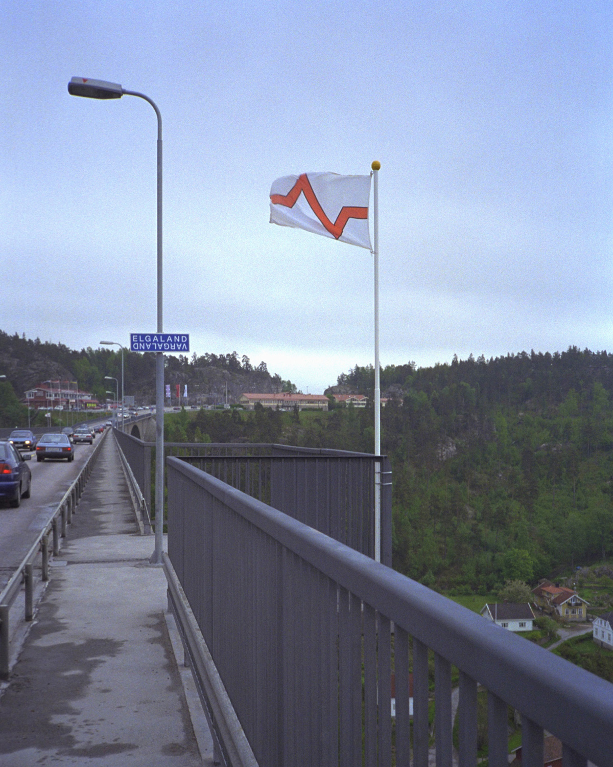 A photograph of a flag on the edge of a highway that marks Elgaland-Vargaland’s physical territory, Svinesund, on the border between Sweden and Norway.