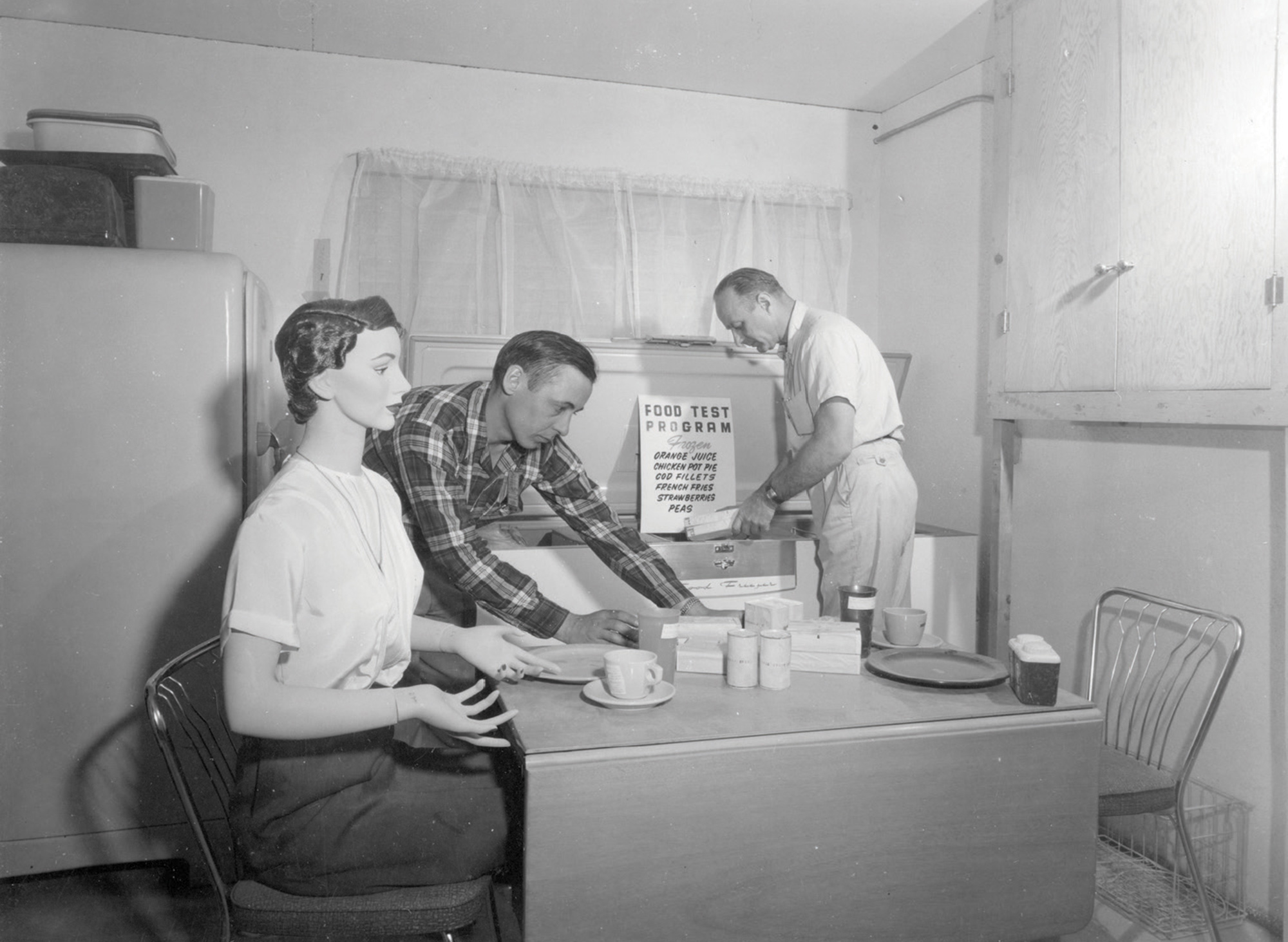 A photograph from Operation Cue depicting two workers installing a female test dummy at a kitchen table.