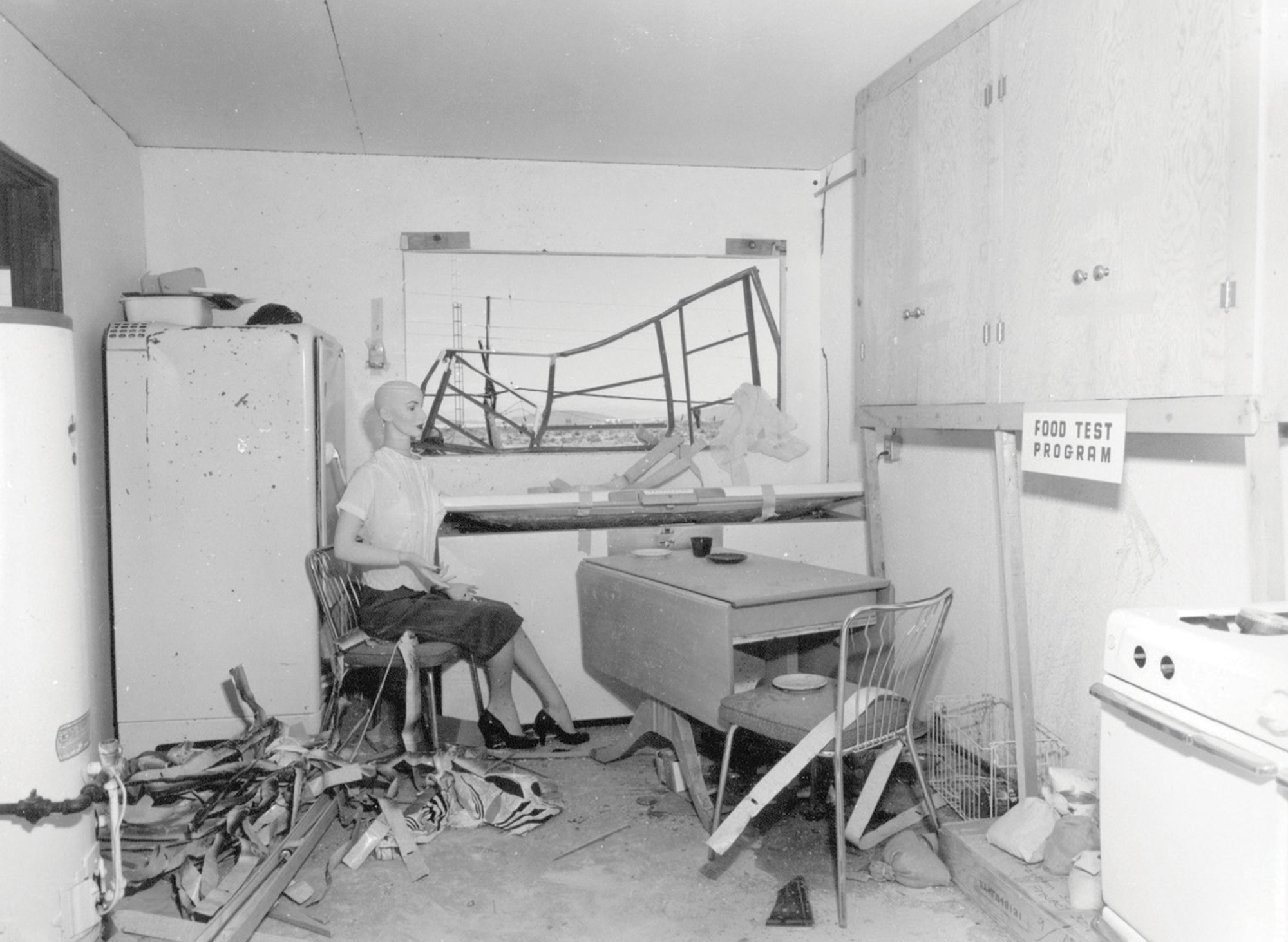 A photograph from Operation Cue depicting a damaged female dummy in the kitchen. 