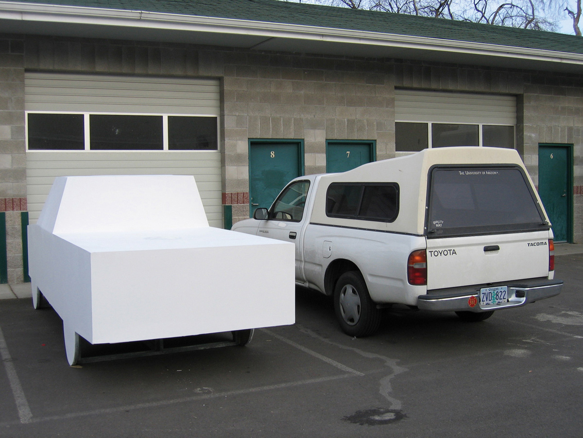 A photograph showing two vehicles parked next to each other, one a normal Toyota pickup truck and the other a lifesized wooden cutout of a pickup truck.