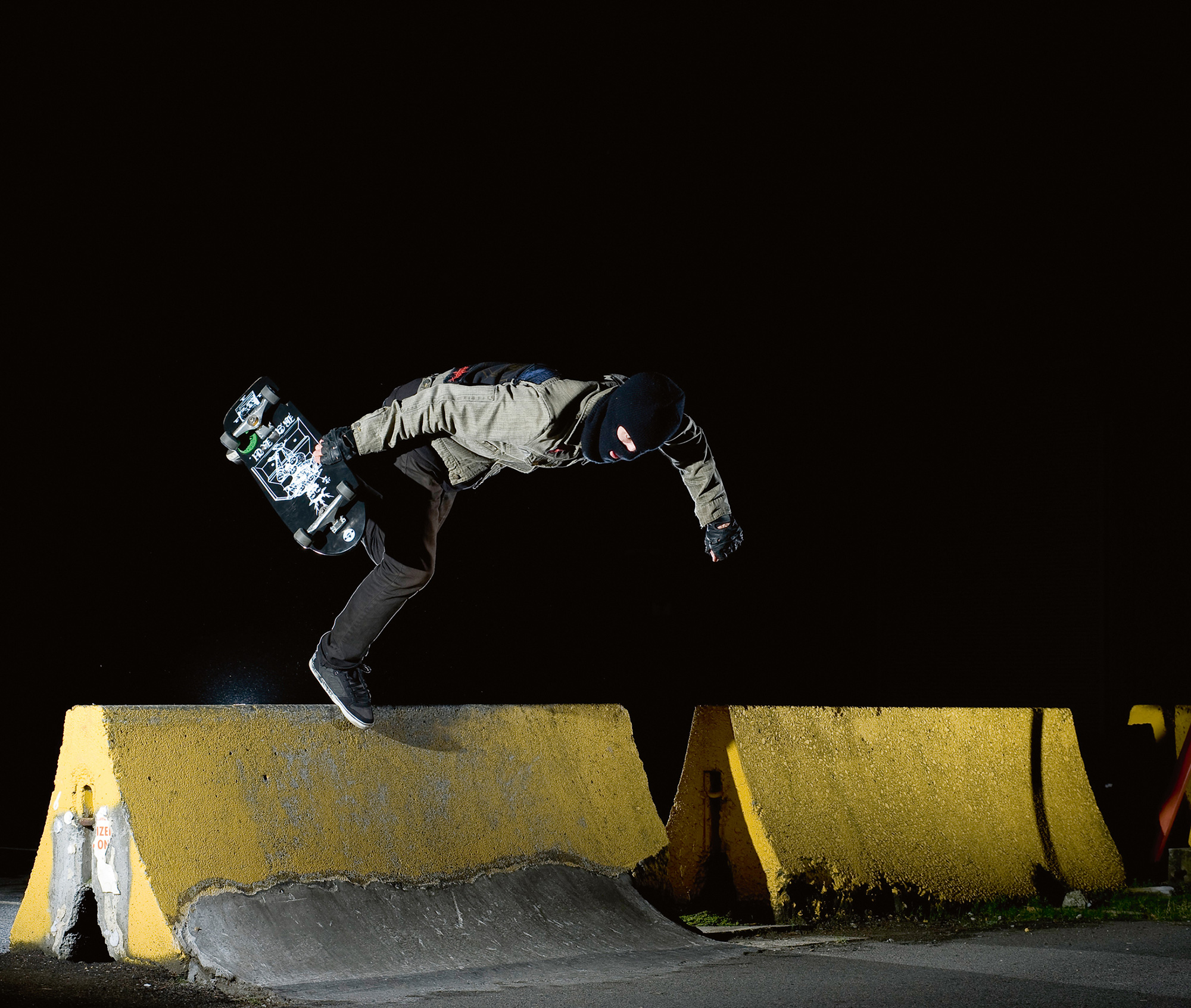 A photograph of the skateboarder known as ”Deer Man of Dark Woods” using a Jersey barrier to do tricks. 