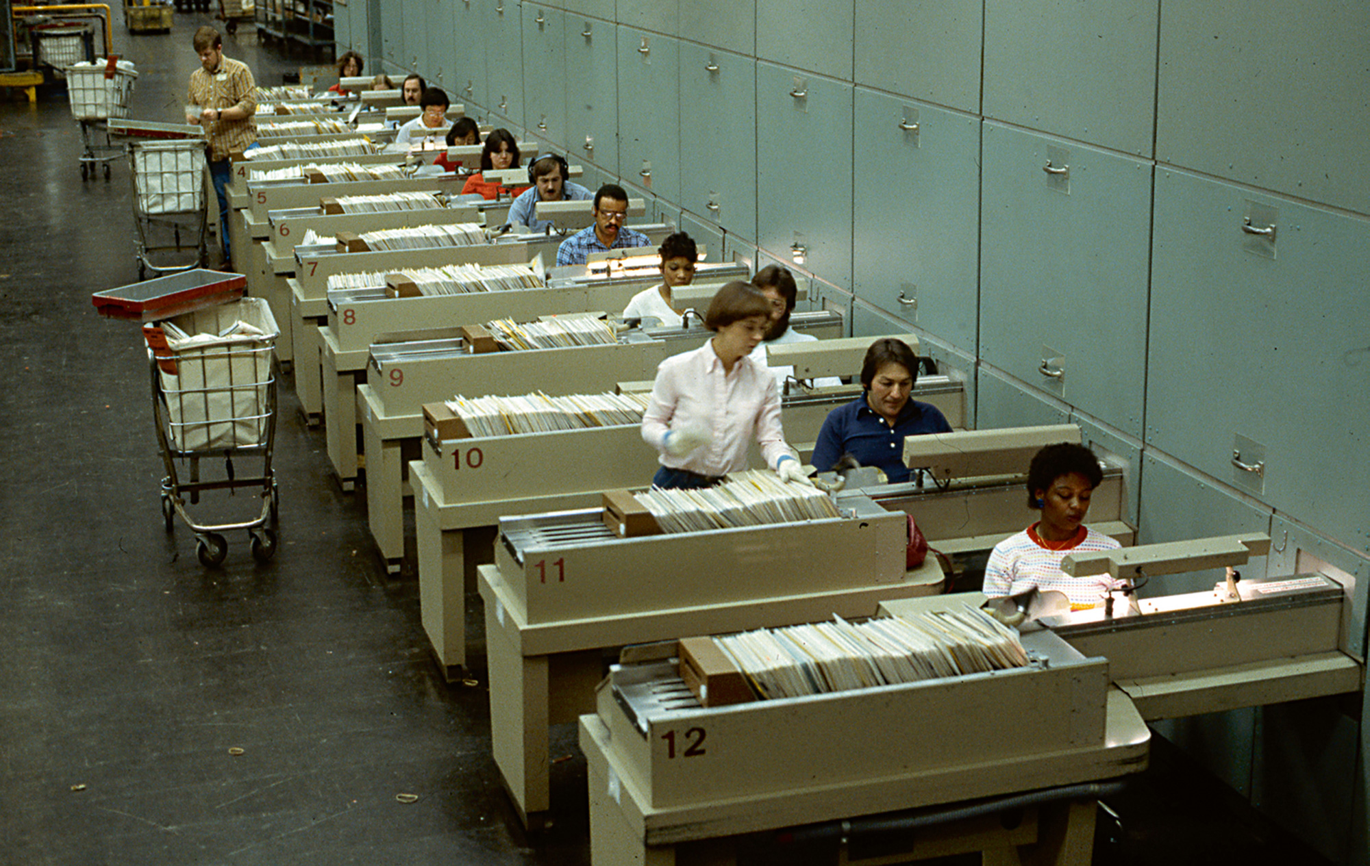 A circa nineteen seventies photograph of postal employees at Multi-Position Letter Sorting Machines processing envelopes using the zip code on each.