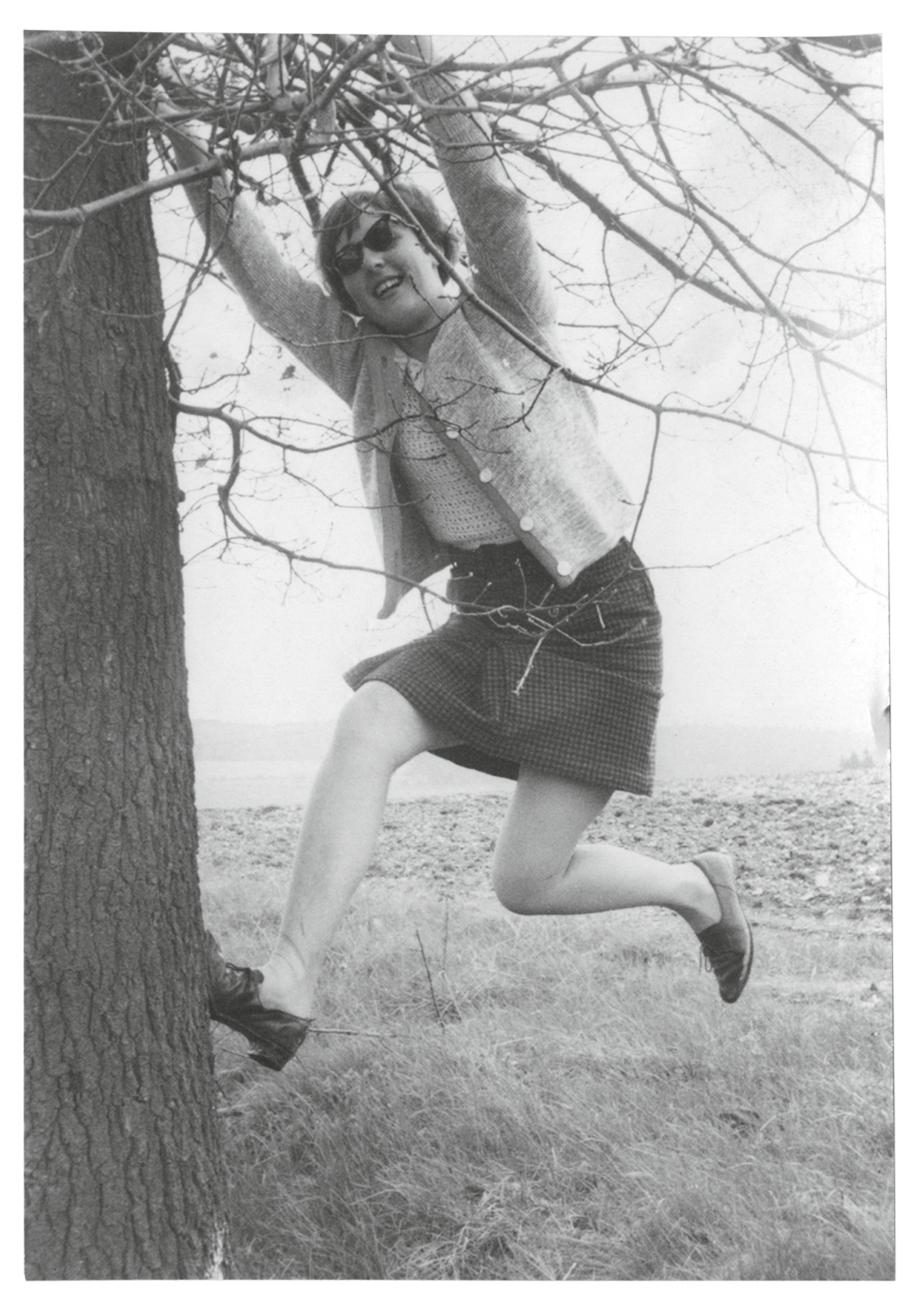 A photograph of a woman in a skirt and cardigan hanging from a tree branch. 