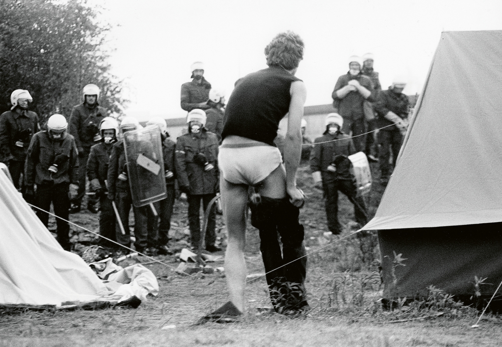 A photograph of a man getting dressed outside his tent as West German police surround the encampment built by protesters on the Lenné Triangle in the summer of nineteen eighty-eight. 