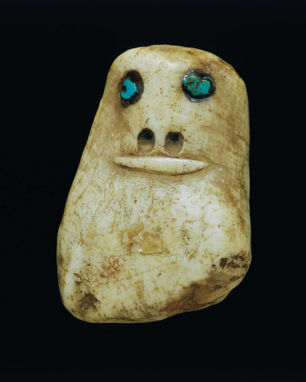 An undated photograph of a Native American figurine from Nambe Pueblo, New Mexico. It is carved to represent a face with round, turquiose eyes.
