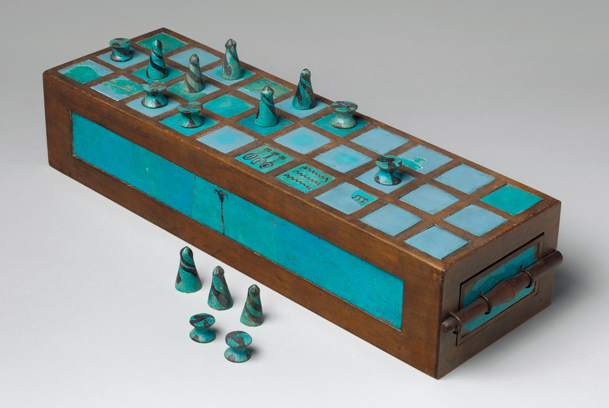 A photograph of an engraved wood game box and small pieces for the ancient Egyptian game of senet. The faience inlays and playing pieces of this game box were discovered in a tomb at Abydos. The Metropolitan Museum of Art estimates that these artifacts were made between fifteen fifty BC and twelve ninety five BC; the box itself is a reconstruction.