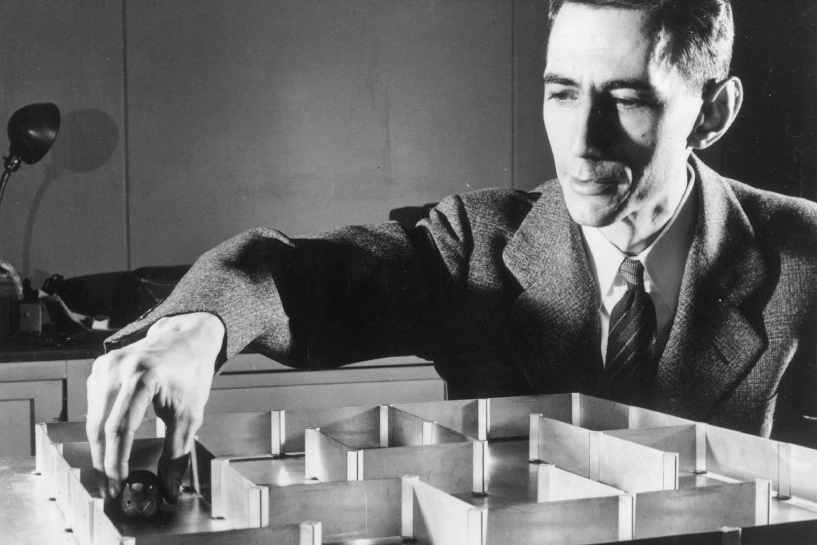 A photograph of mathematician Claude Shannon guiding his labyrinth-traversing mouse Theseus, invented in nineteen fifty, through a tabletop maze.