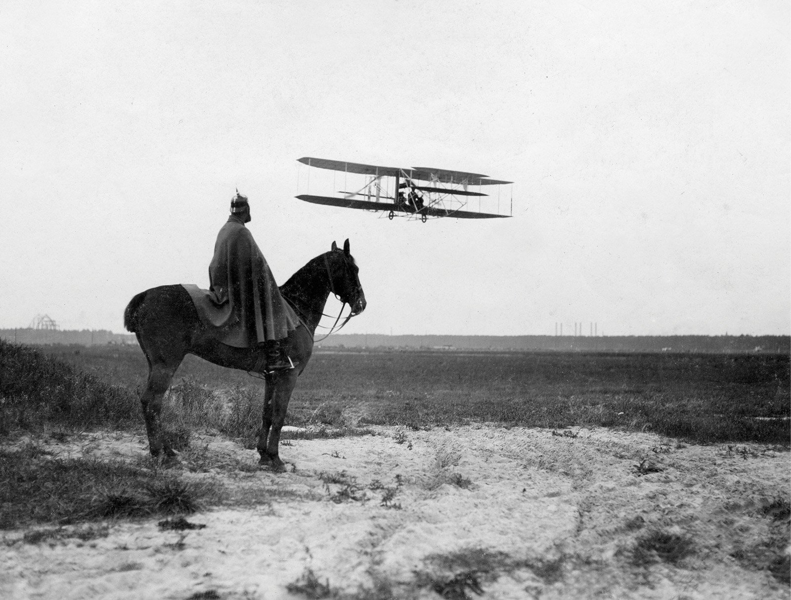 A nineteen ten photograph on the front of a postcard, depicting an indigenous man atop a horse gazing at a plane as it prepares to land at Johannisthal airfield in Berlin. 
