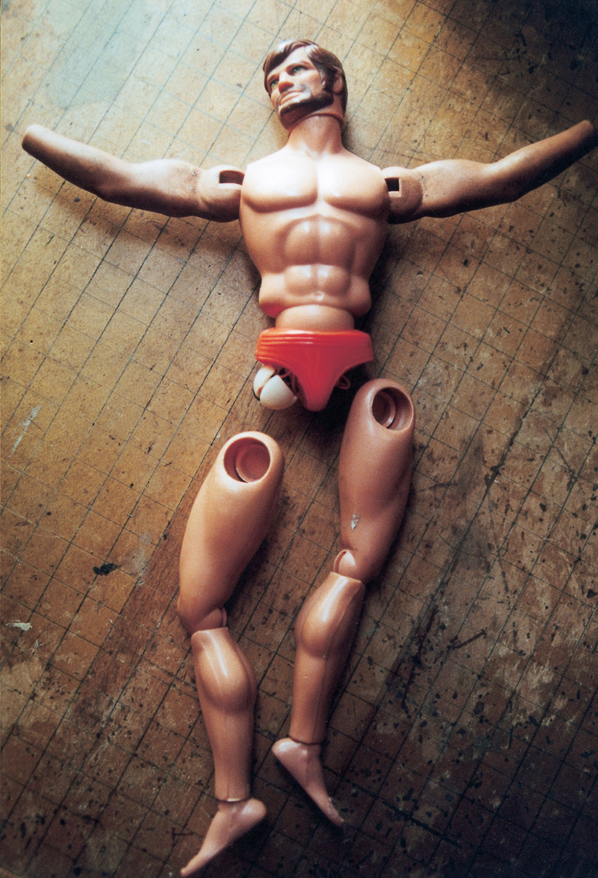 A photograph depicting a bearded male doll with his legs detatched named Josh McBig. The caption reads: The Doll Games’ archetypical “manly man” with action arm, originally used to chop a plastic log (now lost). His name seems to gently parody his original identity as brawny lumberjack, “Big Josh.” In spite of his muscled torso, moustache, and the mature bulge in his plastic underwear, all of which marked him as masculine “other” to the youthful androgynes Laurie and Aina, and no doubt to the young Jacksons, Josh had a vulnerable quality. His legs dangled weakly from his loosely jointed hips, and later began to loosen and fall off, as did his hands and even eventually his left foot, making him a source of comedy, especially in the late farces, but also transforming him, in the end, into what we can only see as the Doll Games’ sorrowful, suffering Christ.
