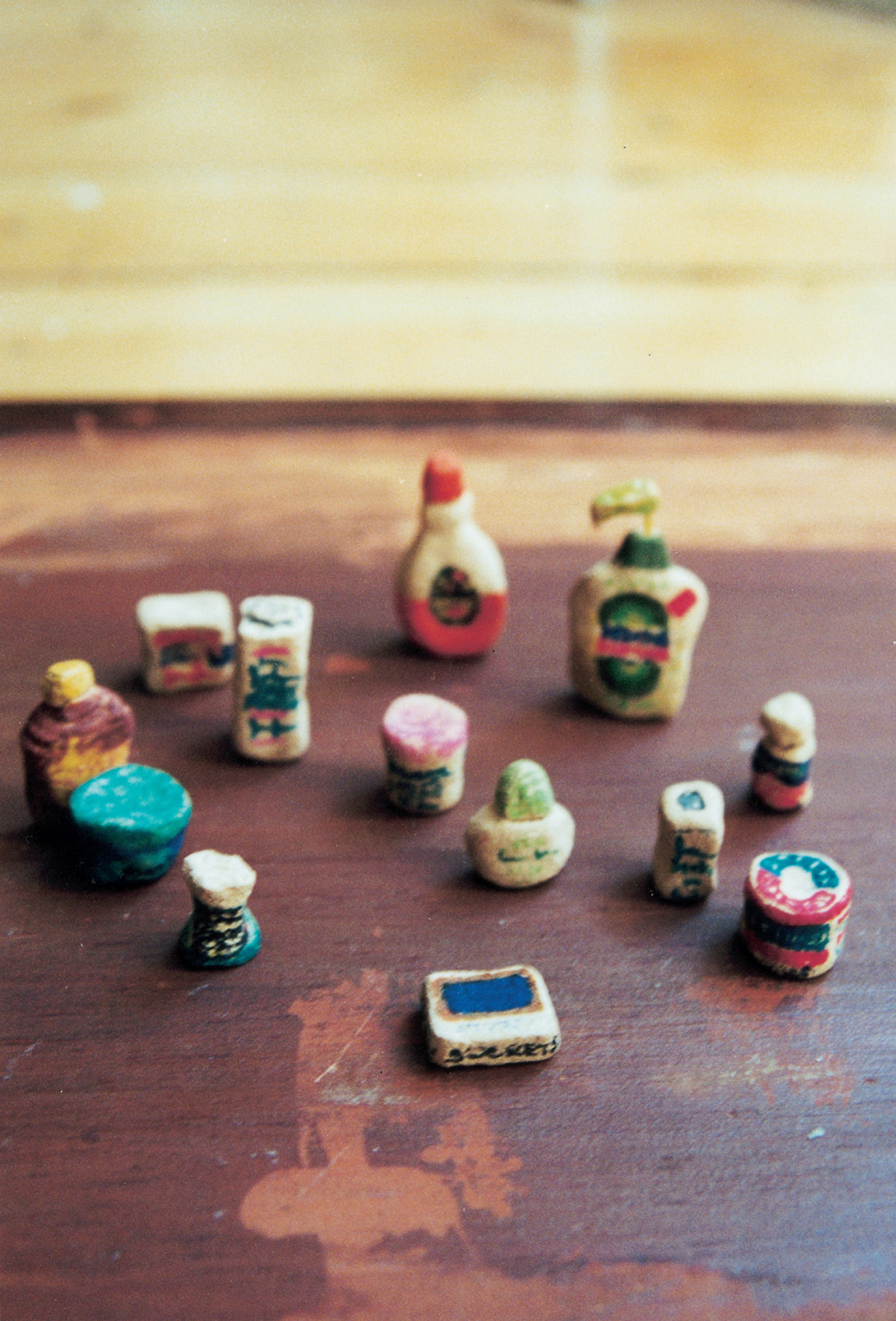 A photograph depicting small modeled objects. The caption reads: Bathroom commodities in molded “plastic wood” decorated with magic marker to resemble name brands, including Desitin lotion (in pump dispenser), Stridex medicated pads, Secret deodorant, Noxzema, Band-Aids, Johnson’s Baby Cream, Suave Shampoo, Johnson’s Baby Powder, Coppertone, Sucrets, Johnson’s Dental Floss, [brand illegible] nasal spray, Buffered aspirin. Emblematic of the way the Doll Games consumed the larger culture, transforming and reconfiguring it for its own purposes, these jewel-like miniatures, none more than half an inch tall, are the products of thoroughly American dreamers.
