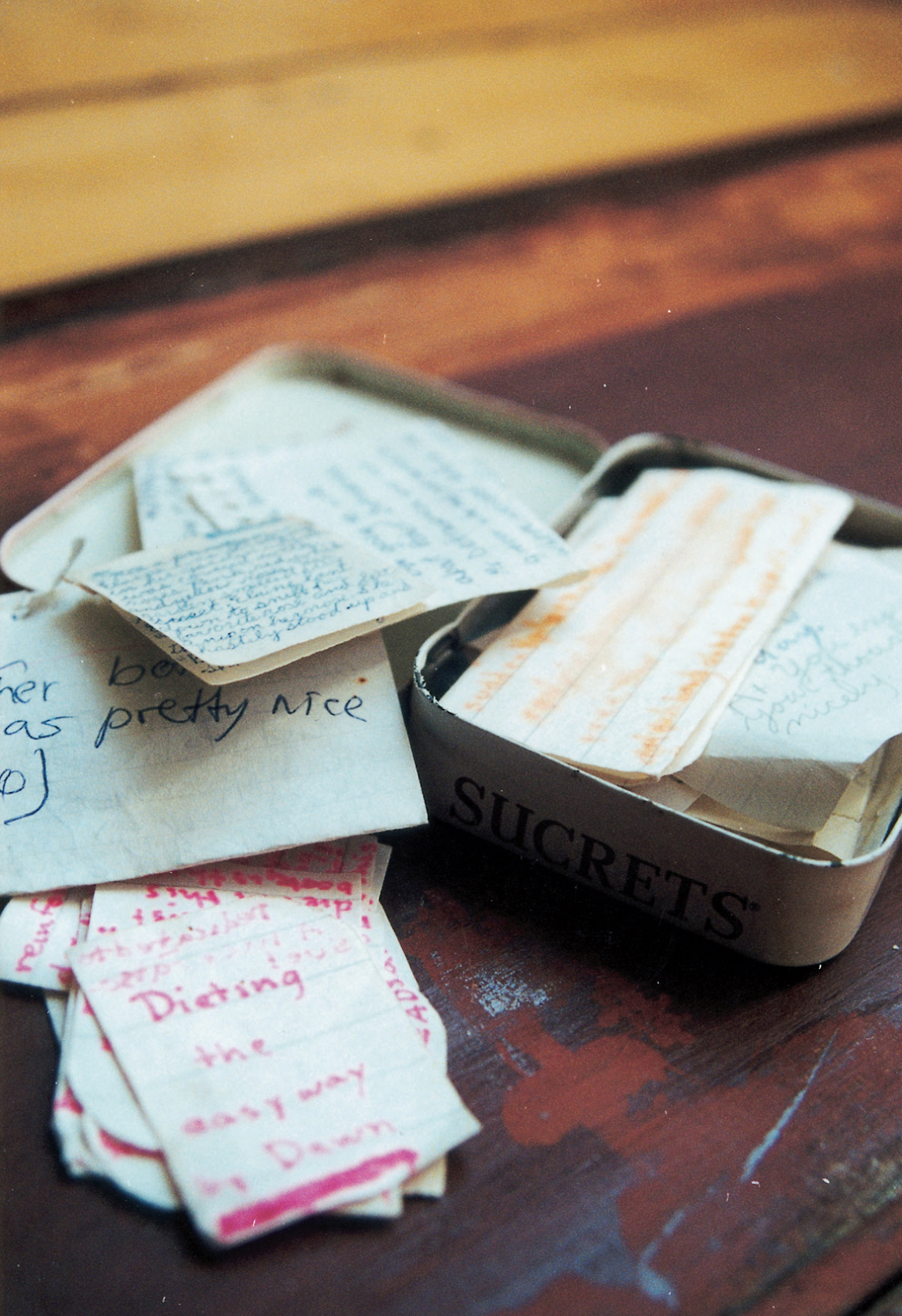 A photograph depicting a small metal box containing scraps of paper. The caption reads: Tin “SUCRETS” box containing the assembled writings of the dolls in various styles and formats, including literary efforts (Dawn’s “A true-life romance”, Harvey’s “Moments with Mara” and “Parakeet—A flash”), self-help publications (“Dieting the Easy Way, by Dawn,” “Madame Dotrovthnile’s Hairdressing Book”), and romantic ephemera from Dawn’s busy love life. Sucrets, a throat lozenge, was popular with the orally fixated Jacksons for its candy-like sweetness. Note that the only written texts generated within the primarily oral tradition of the Doll Games are preserved in what one might call a “voice box”! The resemblance of Sucrets to secrets will not escape the attention of the careful reader.